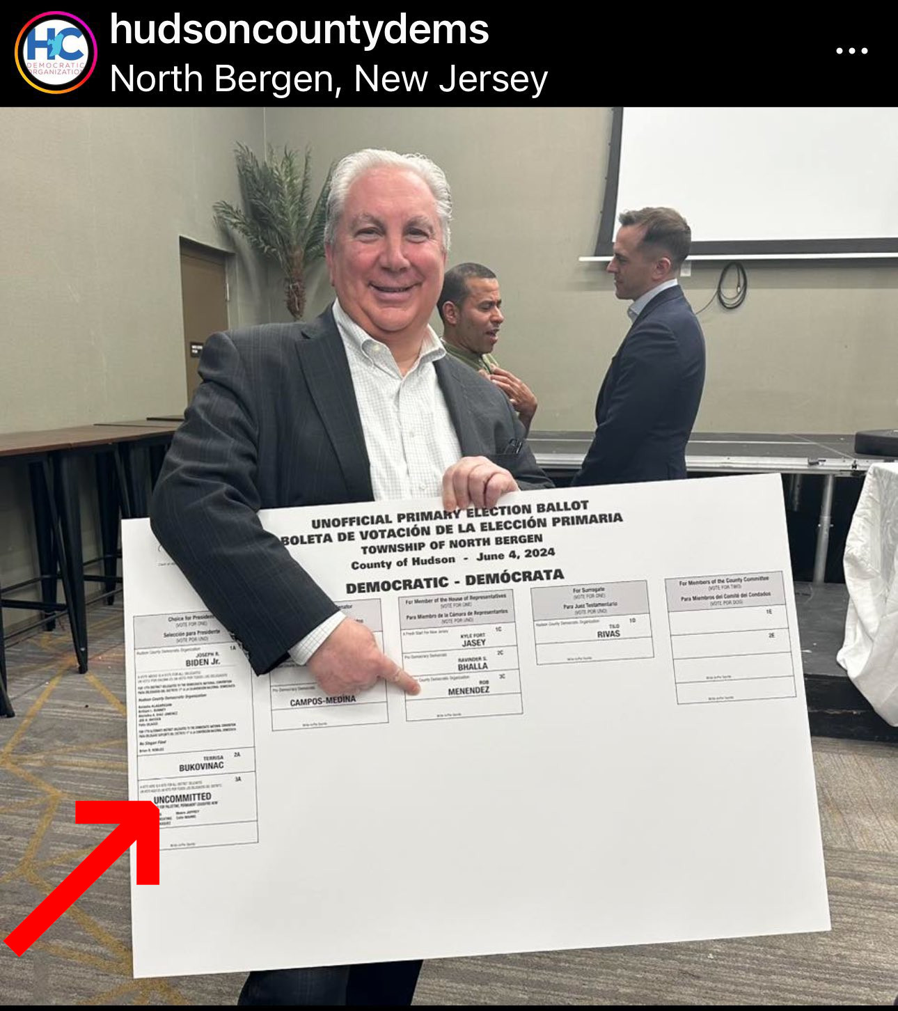 Image of Hudson County Dems on Instagram. The Hudson County Democratic
Organization Chair Anthony Vainieri, a short white man with a receding hairline
is wearing a suit and holding a large white poster which is the ballot mock-up
for Hudson County, with a finger pointing at a candidate. A Red Arrow is
pointing to Uncommitted.